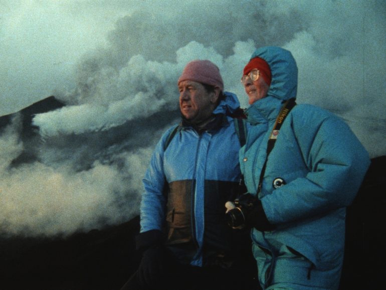 Katia y Maurice Krafft contemplan un volcán en la distancia mientras columnas de ceniza, vapor y gases ondean tras ellos.